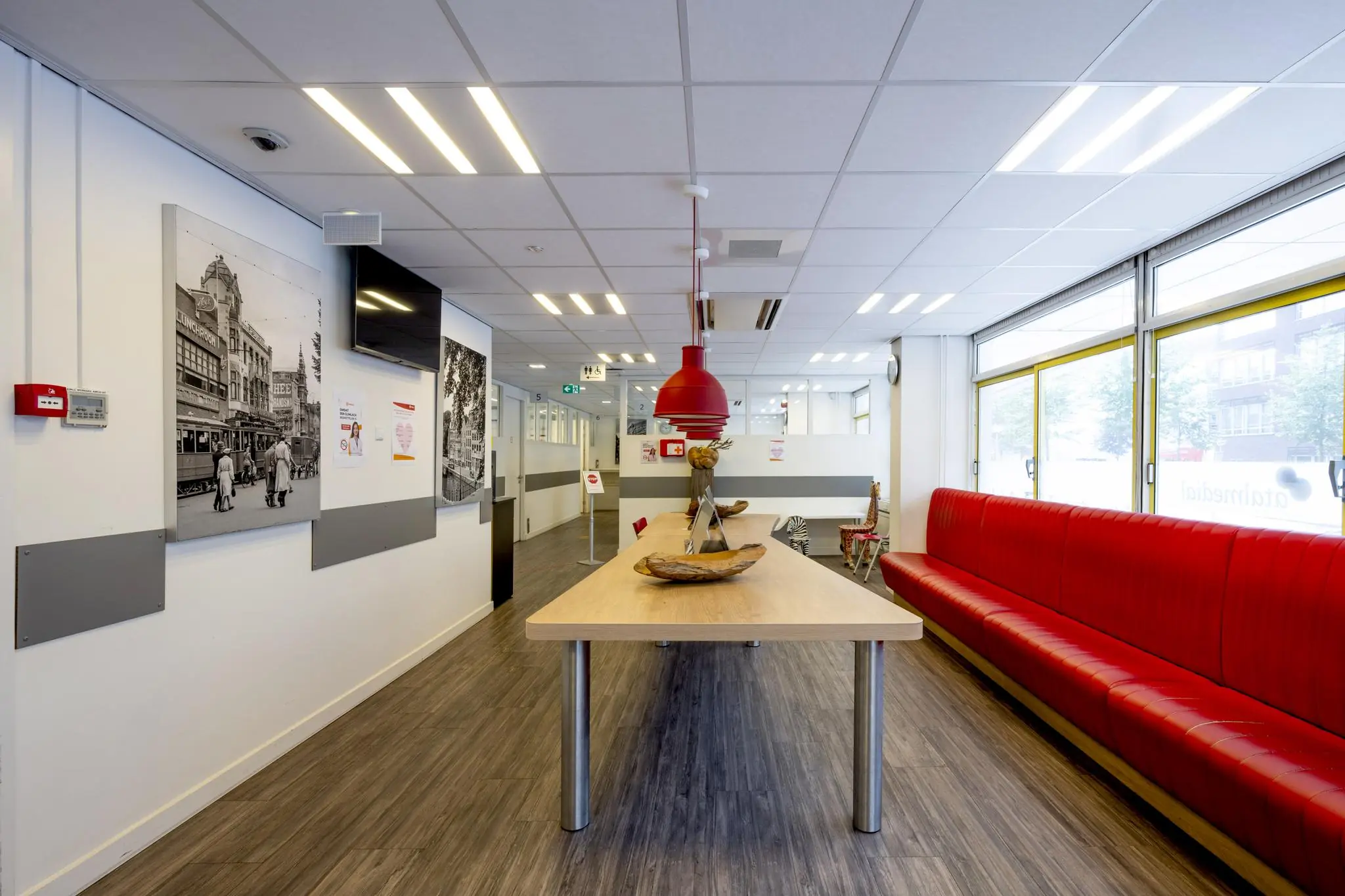 Moderne wachtruimte aan de Jan Tooropstraat met een lange tafel, rode bank en zwart-witfoto’s aan de muur.